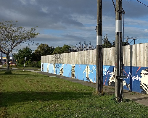 A wall with a blue mural on it with grass and power poles in front.